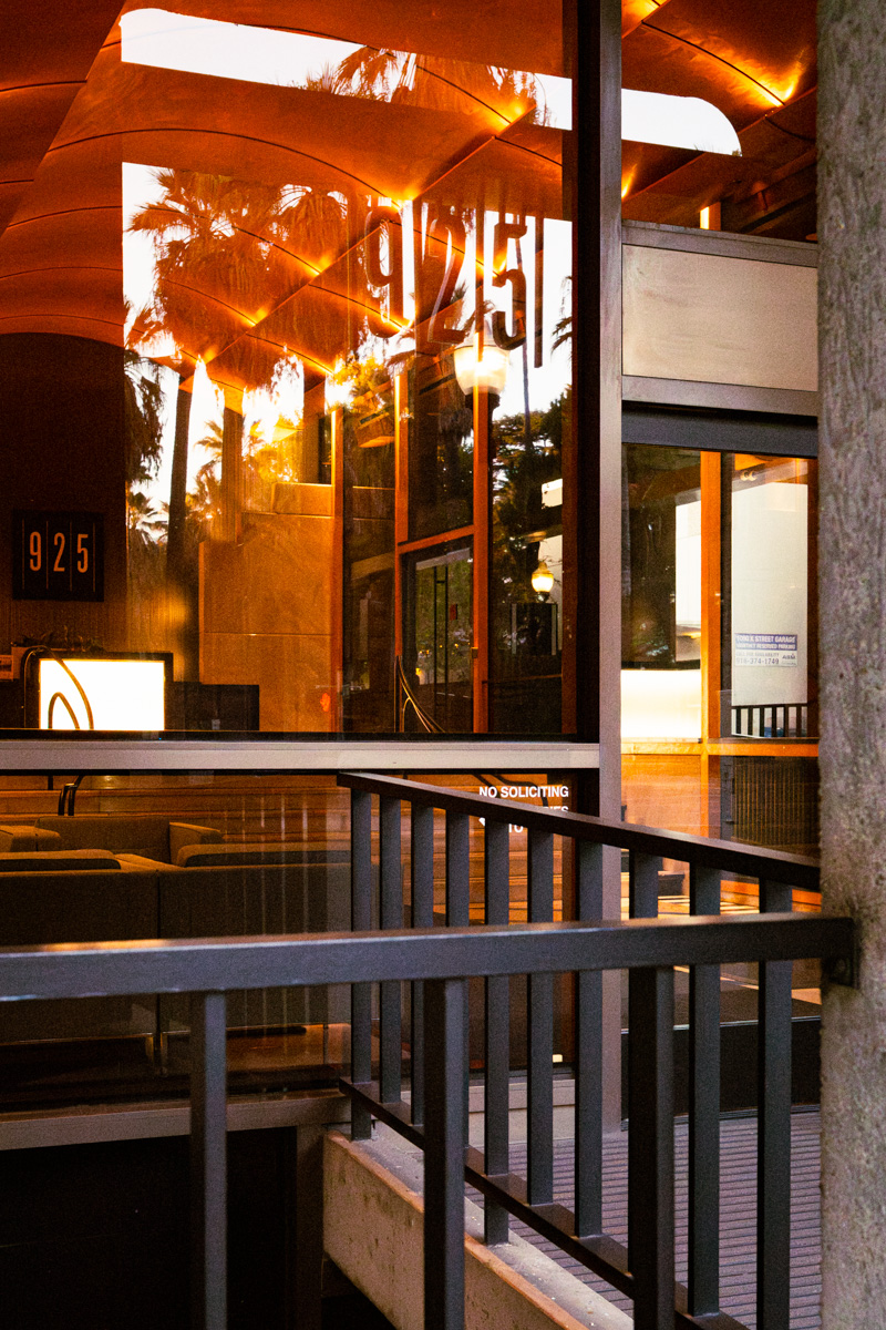 Glass lobby reflecting palm trees and warm light, with a 925 sign and a railing in the foreground