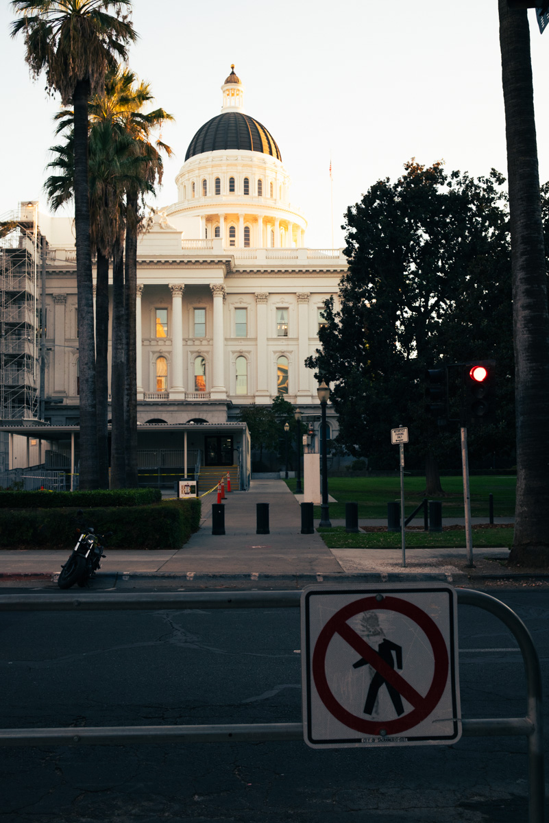 State Capitol dome at dusk behind palm trees, with a no-pedestrian sign in the foreground